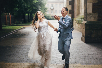Naklejka premium Stylish happy bride and groom running on background of old church in rainy street. Provence wedding. Beautiful emotional wedding couple smiling and having fun in rain in european city.