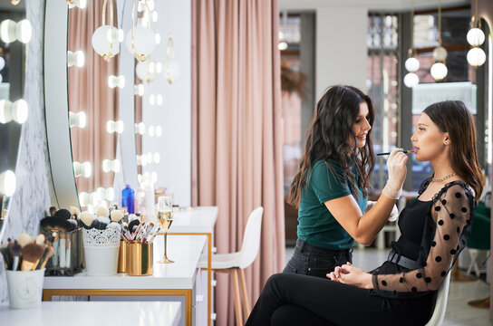 Female Beauty Specialist In Sterile Gloves Applying Lipstick On Client Lips With Cosmetic Brush. Stylish Woman Sitting At Dressing Table While Makeup Artist Doing Professional Makeup In Visage Studio.