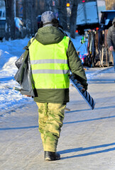 A utility worker with a shovel walks along the sidewalk on a winter day