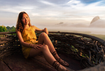 Young Asian beautiful woman traveling alone, sitting on a nest chair to see the sunrise in the morning, summer holiday trip to relax and see the beautiful island in the ocean. Journey voyage