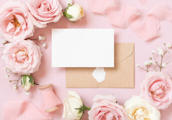 Obraz na płótnie Canvas Card with envelope between pink roses and pink silk ribbons on pink top view, wedding mockup