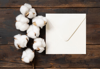 Blank square envelope with cotton flowers top view on dark brown wood, wedding mockup