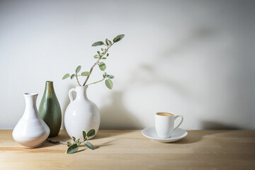 Pale still life with three vases in white and green, a sage leaf branch with shadow and a coffee cup, wooden table against a gray wall, take a break with simple home decor, copy space