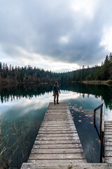 Caucasian young man with a beanie holding camera standing on a pier near a lake with forest background.