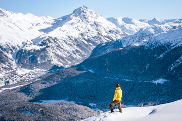 Caucasian man standing on top of a hill looking at winter snow mountain landscape and forests on a sunny day around St. Moritz, Switzerland.