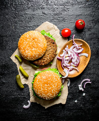 Burgers on paper with chopped onion in bowl and tomatoes.