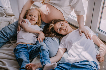 Cropped shoot of two cheerfull sisters in blue jeans and white t-shirts laying on the floor close to pregnant moms belly looking at camera. Happy family at home playing together. Pregnancy concept.