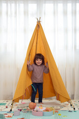 Smiling female toddler standing inside a teepee and looking at camera in a playroom. © Esther Pueyo