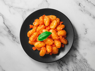 A plate of POTATO GNOCCHI in tomato sauce and a basil leaf on a white marble table. One object top view.