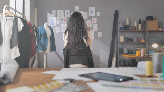 Young female fashion designer looking at clothes sketches on the wall in search for inspiration,back view of creative dressmaker sitting on a chair in studio looking for new idea,startup concept