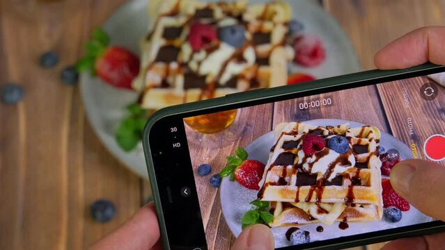 Food blogger recording a video of sweet dessert, woman using smartphone, taking a photo of pile of soft fluffy waffles served with fresh berries and chocolate sauce, food photography, 4k video footage