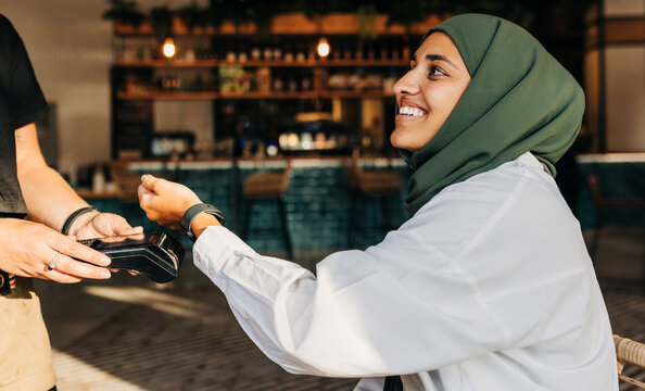 Happy Muslim woman paying her bill using her smartwatch in a cafe