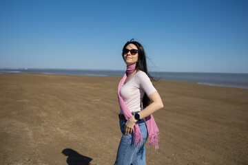 long-haired brunette girl in jeans and a pink scarf walks along the shore
