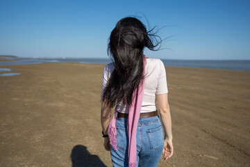 long-haired brunette girl in jeans and a pink scarf walks along the shore