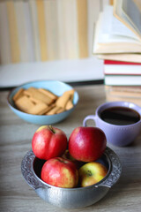 Stack of books, e-reader, reading glasses, bowl of biscuits, apples and cup of tea on the table. Bookshelf in the background. Selective focus.