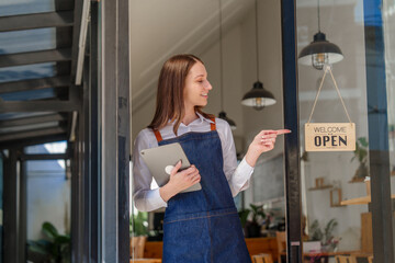 Fototapeta na wymiar Young friendly female pretty caucasian coffee shop woman owner in apron with open sign, small business owners smiling and service take away orders from food delivery application on tablet.