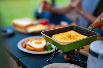 Asian couple cooking food fried egg and bread with sausage for breakfast near the tent at camp. Man and woman enjoy outdoor lifestyle travel nature and camping together on summer holiday vacation.