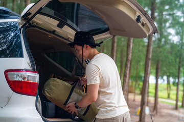 Asian man enjoy outdoor lifestyle on road trip hiking and camping on summer holiday travel vacation. Handsome guy taking off camping supplies and equipment from car trunk at natural park forest.