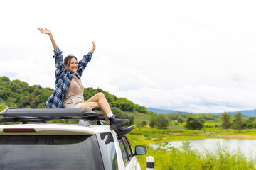 Young Asian woman enjoy and fun outdoor lifestyle road trip and camping in forest mountain on summer travel vacation. Happy attractive girl sitting on car roof rack by the lake with arm raised.