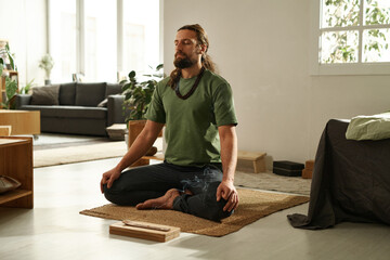 Young man sitting in lotus position and meditating with his eyes closed at home in the morning