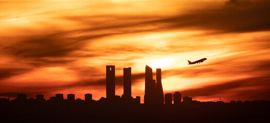 The sun silhouettes a plane as it flies over the skyscrapers of Madrid's skyline during sunset with a panoramic view - obrazy, fototapety, plakaty