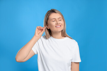 Young woman cleaning ear with cotton swab on light blue background