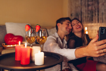 Valentines Day loving couple taking selfie on phone by wine glasses with strawberries having romantic dinner at home