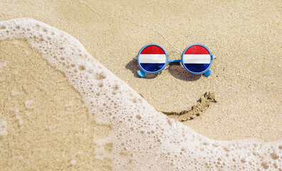 Sunglasses with flag of the Netherlands on a sandy beach. Nearby is a sea lightning and a painted smile.