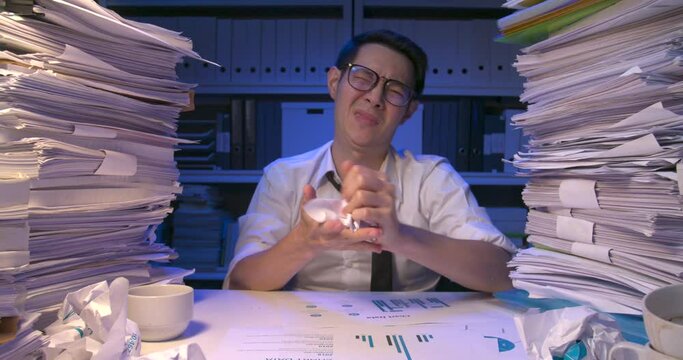 Stressed asian business man is sitting at desk covered with stack of paperwork while working hard at late night. Frustrated alone male is thinking then being stuck, crumpling paper and throws it away