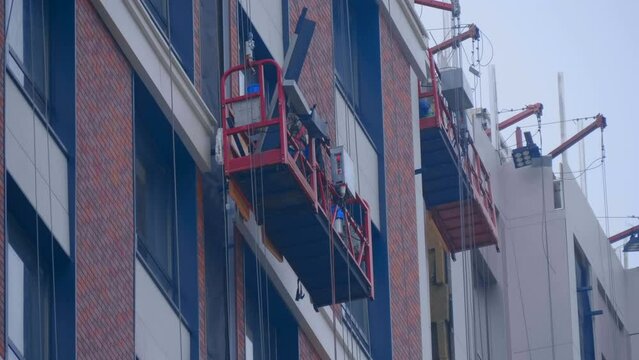 At a construction site, builders in a cradle on cables fastens decorative cladding panels on the wall of a building under construction