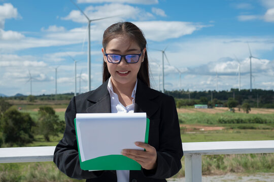 person with folder. businesswoman with clip broad. Asian woman in white helmet working with digital tablet at renewable energy farm. Female inspector controlling functioning of wind turbines outdoors.