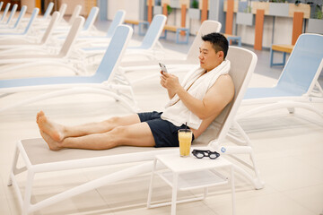 Asian man sitting on a sunbed in indoors swimming pool and using a phone