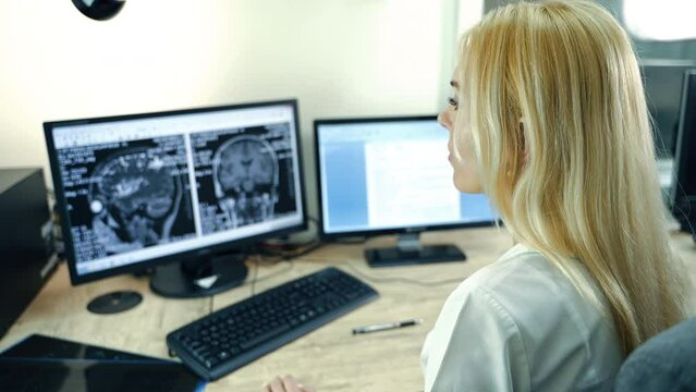 Rear view of a female lab technician sitting in front of computers. Medic looks at the MRI scan of head.