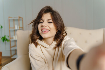 Naklejka premium Pretty young asian female with big smile sitting at living room. She having fun taking light cheerful selfie on blurred background
