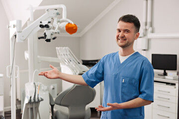 Handsome young dentist in white coat is looking at camera and showing his office