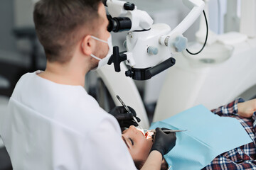 Skilled user. Handsome young male dentist looking into the lens of a professional microscope and treating teeth.