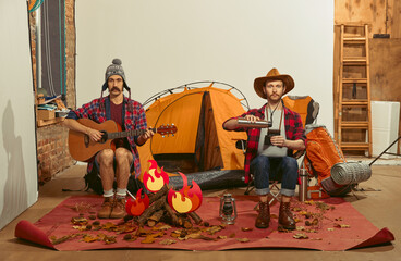 Playing guitar, drinking tea. Two men imagining, recreating camping activity indoors with necessary...
