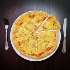 Delicious fresh round cheese pizza with fish, lemon on black table, on white plate, fork, knife nearby, top view