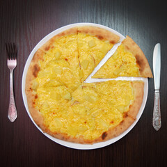 Delicious fresh round cheese pizza with pear, pineapples on black table, surface, on white plate, fork and knife nearby, top view