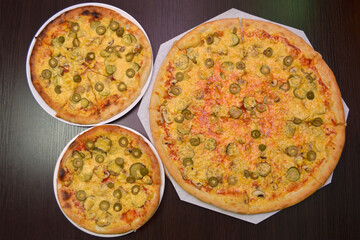 Three delicious fresh round cheese pizzas with mushrooms, pickles, olives on a black table, surface, on a white plates, three different sizes for serving in a pizzeria