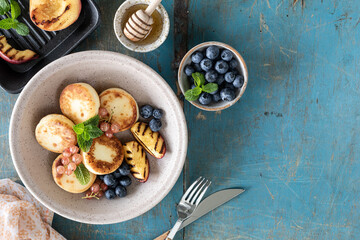 Fototapeta na wymiar Cottage cheese pancakes, cheesecakes, ricotta fritters with fresh blueberries, currants and peaches on a plate. Healthy and delicious breakfast for the holiday. Blue wooden background