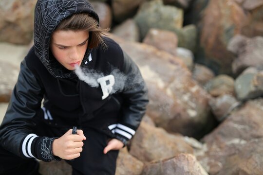 a guy of thirteen years old, holds a vape in his hands. A teenager smokes a disposable electronic cigarette while sitting by the sea