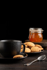 Close-up of cup of coffee, plate with cookies, spoon and jar of jam on dark table, black background, vertical, with copy space