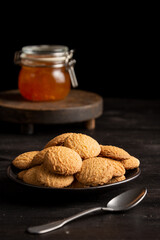 Close-up of plate with cookies, spoon and jar of jam on dark table, selective focus, black background, vertical, with copy space