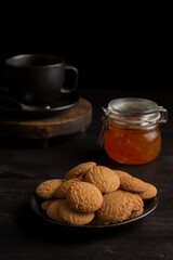 Top view of plate with cookies, cup of coffee and jar of jam on dark table, selective focus, black background, vertical, with copy space