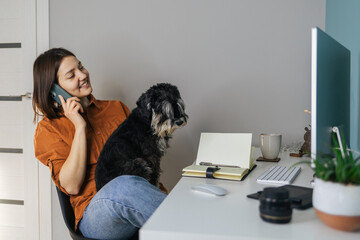 Young caucasian lady speaks on the phone and works at the computer with her little black dog. Freelance concept