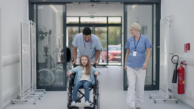 Young caregiver pushing little girl on wheelchair at hospital corridor, having fun together.