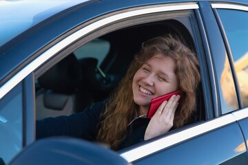 happy girl, young woman is sitting in her car, automobile, enjoying driving. Joyful lady in auto using, talking on cell mobile phone, calling on smartphone. Dangerous situation, careless driver