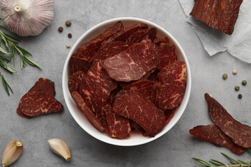 Delicious beef jerky in bowl and seasoning on light gray table, flat lay