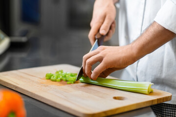 Obraz na płótnie Canvas cooking food, profession and people concept - close up of male chef with knife chopping celery on cutting board at restaurant kitchen
